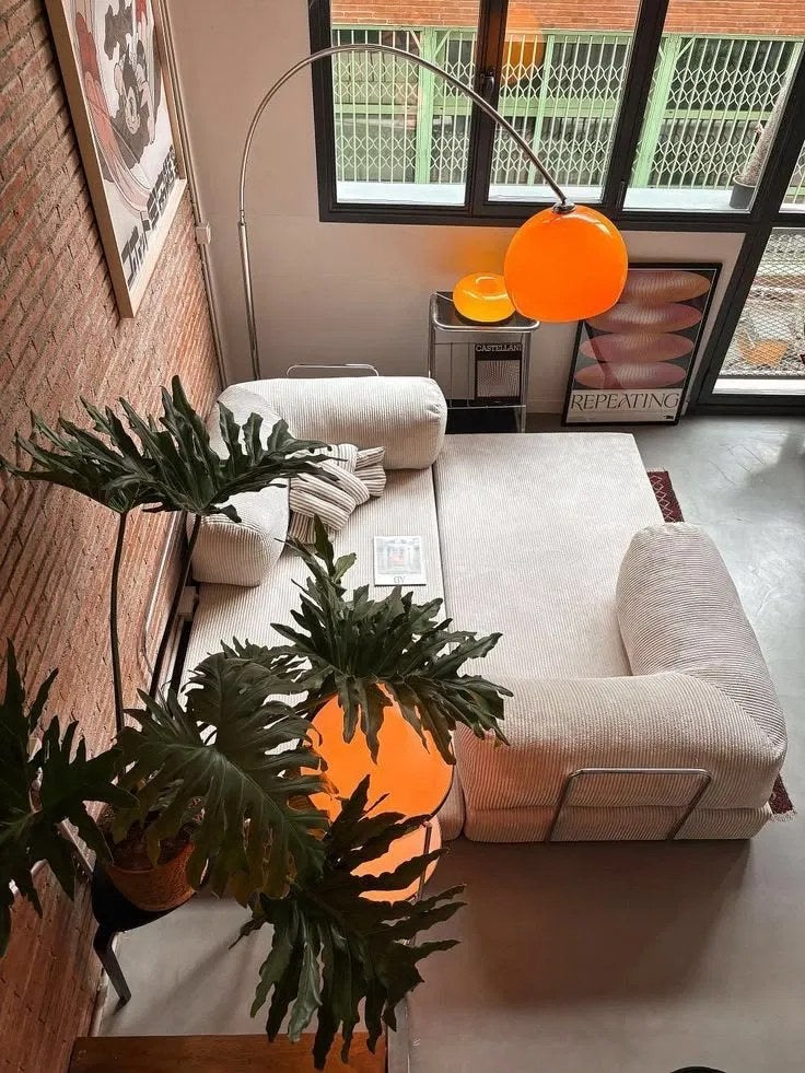 Vue plongeante d’un loft : canapé en velours côtelé clair, mur de brique, plantes, lampe arc orange